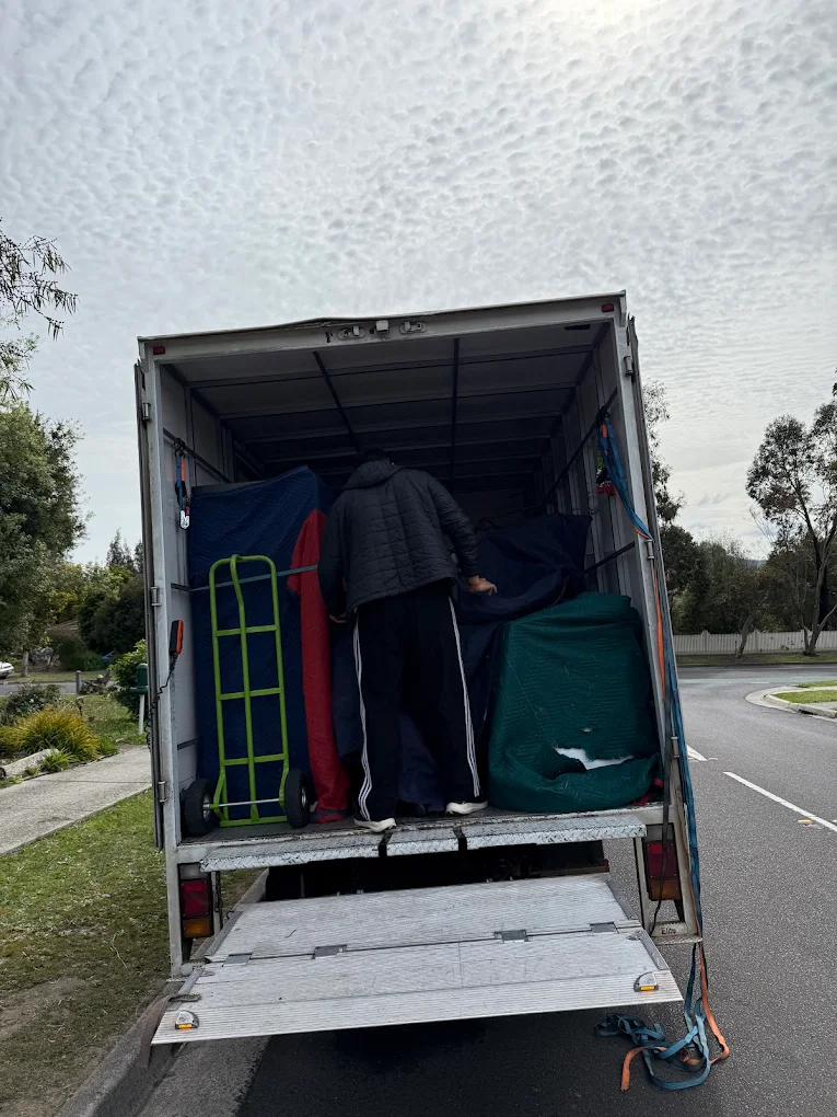 Big Stuff Movers team loading a truck with furniture wrapped in moving blankets and a green hand trolley on a suburban street.
